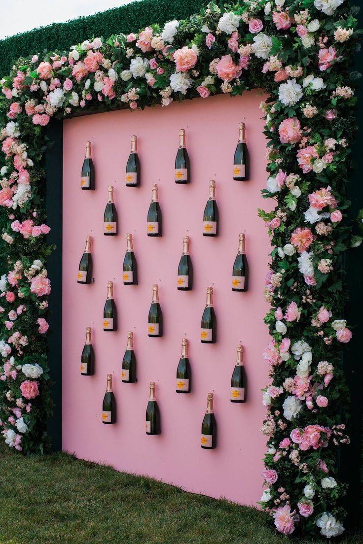 Painel instagramável em evento da Veuve Clicquot com fundo rosa e garrafas fixadas horizontalmente com moldura floral